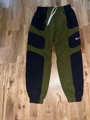 Nike gröna och svarta cargopants - Säljer ett par feta cargopants från Nike i olivgrönt och svart med vita snören i midjan. Byxorna har elastiska muddar vid bensluten, dragkedjor på fickorna och den klassiska Nike-loggan på benet. Perfekt för dig som gillar streetwear och sportig stil.