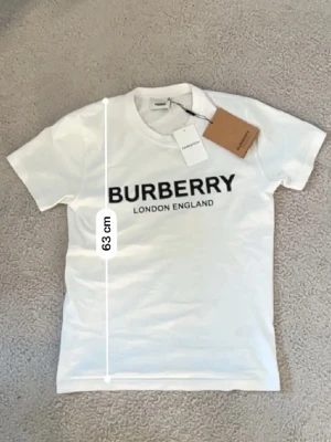 Vit Burberry t-shirt med tryck - Vit t-shirt från Burberry med klassisk svart logotyp 'BURBERRY LONDON ENGLAND' tryckt på bröstet. Rund halsringning och korta ärmar. Tillverkad i mjuk bomull för en skön känsla. Perfekt för dig som gillar stilrena designer och ikoniska märken.Aktiv 24/7✅ Skickas inom 24h📦🚚 Kan gå ner i hugget 😃✅ Hör av er via frågor⁉️