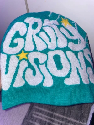 Två stickade mössor med tryck - Säljer två stickade mössor, en i turkos med vit text och gula stjärnor, och en i vit med svart text och röda stjärnor. Båda har coolt 'GRIM VISIONS'-tryck på ena sidan och spindelnätsmönster på andra sidan. Perfekt accessoar för dig som vill sticka ut. Finns i Göteborg att hämtas. Passar alla huvudstorlek. 50kr per mössa!