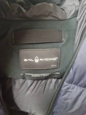 Mörkblå Sail Racing dunjacka S - Säljer en mörkblå dunjacka från Sail Racing i storlek S. Jackan har en puffad design, huva och dragkedja framtill. Logotypen syns både på bröstet och på ryggen. Materialet är syntet och dun, perfekt för kalla dagar.