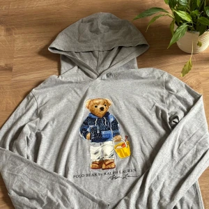 Grå hoodie Polo Bear Ralph Lauren XL - Grå hoodie från Polo Ralph Lauren med ikoniska Polo Bear-trycket framtill. Långärmad och tillverkad i mjuk bomull. Klassisk huva och relaxed fit, perfekt för en chill och stilren look. Perfekt på sommarkvällarna. Storlek XL (170).
