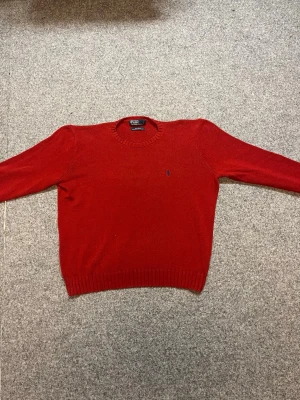 Röd stickad tröja från Polo Ralph Lauren - Säljer en röd stickad tröja från Polo Ralph Lauren med klassisk blå broderad logga på bröstet. Tröjan är tillverkad i 100% bomull, har rund hals och långa ärmar med ribbade muddar vid ärmslut och nederkant. Perfekt för dig som gillar stilren design.äkta