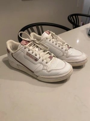 Adidas sneakers i vitt och rosa - Säljer ett par klassiska Adidas sneakers i vitt med rosa detaljer och logga. Skorna har snörning, rund tå och platt sula. Materialet är skinn och de har en låg profil som passar till många olika stilar. Perfekta för dig som gillar retrovibe och snygga färgkombinationer.