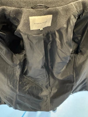 Grå rock från Studio Total, storlek 46 - Snygg grå rock från Studio Total i klassisk modell med knappar framtill och diskreta fickor på sidorna. Jackan har lång ärm och är tillverkad i ullblandning med slät insida. Perfekt för dig som vill ha en stilren och tidlös look.