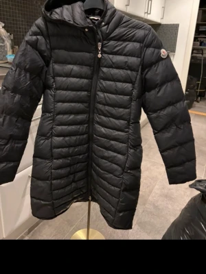 Svart dunjacka från Moncler med huva - Säljer en svart dunjacka från Moncler med huva och snygga horisontella quiltade paneler. Jackan har dragkedja framtill, två sidofickor med dragkedja och Moncler-logga på ärmen. Perfekt för kalla dagar och har en längre passform som täcker bra.