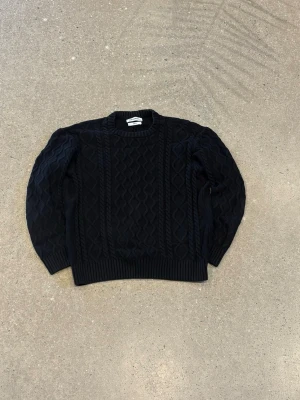 Svart stickad tröja från New Garments - Svart stickad tröja från New Garments i storlek M. Tröjan har ett snyggt kabelstickat mönster framtill och på ärmarna, samt ribbade muddar vid hals, ärmslut och nederkant. Perfekt för dig som gillar klassisk stil med en twist.