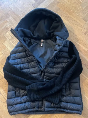 Svart cardigan från Colmar - Säljer en svart pufferjacka från Colmar Originals med stickade svarta ärmar och huva i mjukt material. Jackan har dragkedja framtill och två sidofickor. 9/10 skick, finns inga skador. Skriv om du har en fråga