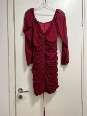 Vinröd kort klänning med snörning - Säljer en vinröd kort klänning med lång ärm och snygg v-ringning. Klänningen har rynkade detaljer längs hela framsidan och snörning nedtill för att justera längden. Materialet känns lätt och har en mjuk finish.