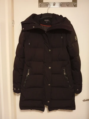 Svart lång pufferjacka från Hollies - Säljer en svart pufferjacka från Hollies med huva och snygga detaljer som dragkedjor och knappar. Jackan är lång, har två fickor med dragkedja och coolt tryck på insidan. Perfekt för kalla vinterdagar och har en stilren look.