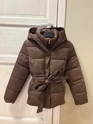 Brun pufferjacka med huva och bälte - Är liten i storlek, passar bättre en i M och kanske L. Helt oanvänd pga storleken. Snygg brun pufferjacka med huva och bälte i midjan. Jackan har quiltad design, dragkedja och knappar framtill. Perfekt för kalla dagar och har en loose passform som ger en chill vibe. Materialet är syntet och jackan är långärmad.