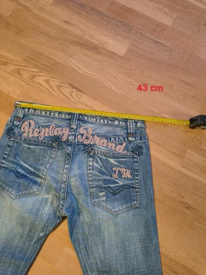 Replay Brand blå jeans med broderi - Säljer ett par blå Replay Brand jeans med coolt broderi i rosa på bakfickorna. Jeansen har klassisk femficksdesign, raka ben och slitna detaljer för en avslappnad vibe. Materialet är jeans och passformen är normal. Perfekt för dig som gillar streetstyle.