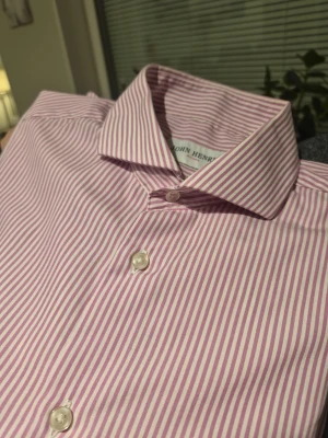 Rosa- och vitrandig skjorta John Henric - Säljer en klassisk långärmad skjorta från John Henric i storlek 40. Skjortan har smala rosa och vita ränder, knappar framtill och på manschetterna samt en snygg krage. Tillverkad i mjuk bomull som känns skön mot huden. Perfekt för dig som gillar stilrena detaljer.