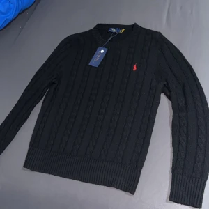 Svart kabelstickad tröja Polo Ralph Lauren - Svart kabelstickad tröja från Polo Ralph Lauren med klassisk rund hals och röd broderad logga på bröstet. Tröjan har ribbade muddar vid ärmslut och nederkant, och är gjord med mjuk bomull. Storlek Small(S).