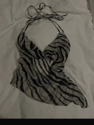Zebramönstrad omlott linnetopp - Säljer en snygg omlott linnetopp med zebramönster i svart och beige. Toppen har tunna knytband i nacken och är ärmlös, vilket ger en cool och trendig look. Perfekt för dig som gillar djurmönster och vill sticka ut.