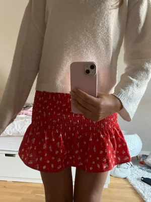 Röd blommig kortkjol  - Supersöt röd kortkjol med vitt blommigt mönster från H&M Divided. Kjolen har smock i midjan och volang nedtill. Tillverkad i mjuk polyester med lite stretch för extra komfort. Perfekt för sommar dagar!