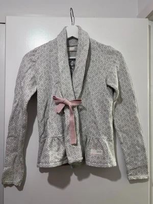 Grå mönstrad kofta med rosa rosett - Supermysig grå kofta från odd molly med diskret mönster och en söt rosa rosett framtill. Koftan har lång ärm, mjukt material och en avslappnad krage. Perfekt för att ge din outfit en gullig touch. 