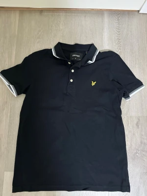 Svart piké från Lyle & Scott - Klassisk svart pikétröja från Lyle & Scott med vitrandiga detaljer på krage och ärmslut. Tröjan har korta ärmar, knappslå framtill och den ikoniska gula logon broderad på bröstet. Perfekt till jeans eller shorts för en clean look.