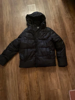 Svart pufferjacka från FSBN L - Säljer en svart pufferjacka från FSBN i storlek L. Jackan har huva, dragkedja framtill och två sidofickor. Den har två små hål men går att sy ihop