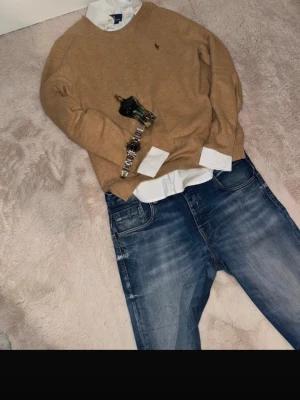 Beige stickad tröja från Ralph Lauren - Snygg beige stickad tröja från Ralph Lauren med klassisk rund hals och diskret logga på bröstet. Perfekt att bära över en skjorta för en clean och stilren look. Tröjan har långa ärmar och ribbade muddar vid ärmslut och nederkant.