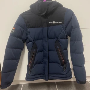 Mörkblå pufferjacka från Sail Racing - Säljer en mörkblå pufferjacka från Sail Racing med svarta detaljer och logga på bröstet. Jackan har två fickor med dragkedja, hög krage och quiltad design. 