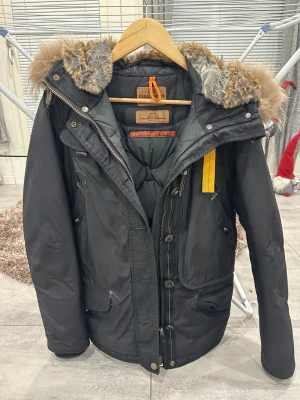 Svart Parajumpers parkajacka med päls - Svart parkajacka från Parajumpers med fet päls på huvan och cool gul detalj på sidan. Jackan har flera fickor, dragkedja och knappar framtill. Slim fit och perfekt för kalla vinterdagar. Märkets patch på ärmen och snygga detaljer gör den riktigt unik.