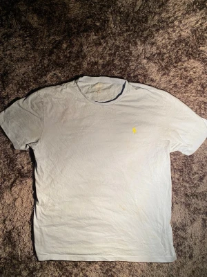 Ljus blå  t-shirt från Polo Ralph Lauren - Ljus blå  t-shirt från Polo Ralph Lauren med klassisk gul broderad logga på bröstet. Modellen är kortärmad och har en rund hals. Tillverkad i mjuk 100% bomull som känns skön mot huden. Perfekt för dig som gillar stilrena och enkla plagg.