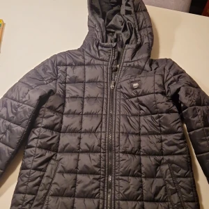 Svart quiltad pufferjacka från Vans - Svart pufferjacka från Vans i storlek Large. Jackan har quiltad design, huva och dragkedja framtill. Snyggt rutmönster över hela jackan och två sidofickor med dragkedja. Perfekt för kalla dagar och enkel att matcha med din stil.
