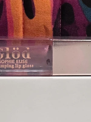 Glöd Plumping Lip Gloss Sophie Elise - Plumping lip gloss från Glöd Sophie Elise i en transparent förpackning med roséguldfärgad text och lock. Ger läpparna extra volym och glans med en lätt rosa ton. Perfekt för dig som vill ha fylligare och glansiga läppar.