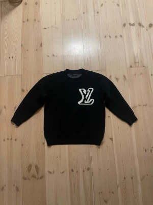Svart stickad tröja Louis Vuitton - Svart stickad tröja från Louis Vuitton med stor vit LV-logga både fram och bak. Rund hals och långa ärmar. Tröjan har en klassisk och clean look med ribbade muddar vid hals, ärmslut och nederkant. Perfekt för dig som gillar exklusiva märken och vill sticka ut.