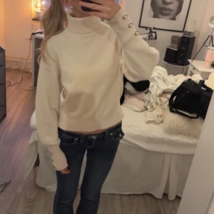 Vit polotröja med pärlknappar - Säljer en snygg vit polotröja med pärlknappar på ärmarna. Tröjan har hög krage, ballongärmar och är lite croppad i modellen. Perfekt för dig som vill ha en stilren och trendig look till hösten eller vintern.