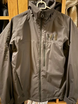 Svart vindjacka Helly Hansen S - Svart vindjacka från Helly Hansen med HH-logga på bröstet och Helly Hansen-tryck på ärmen. Jackan har hög krage, dragkedja framtill och en bröstficka med dragkedja. Materialet är syntet med LifaLoft-isolering för extra värme. Regular fit.