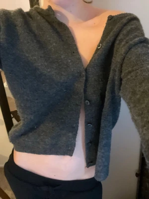 Mörkgrå croppad kofta med knappar - Mysig mörkgrå croppad kofta med långa ärmar och knappar framtill. Tröjan har en loose passform och är stickad i ett mjukt material, perfekt för lager på lager under höst och vinter. Enkel och stilren design som funkar till allt.