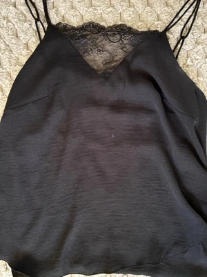 Svart linnetopp med spetsdetalj - Säljer en svart linnetopp med tunna axelband och snygg spets vid v-ringningen. Toppen har en slät yta och är lätt och luftig, perfekt för varma dagar eller att styla lager på lager. Spetsen ger en extra touch av elegans och gör toppen unik. Vet ej märket!