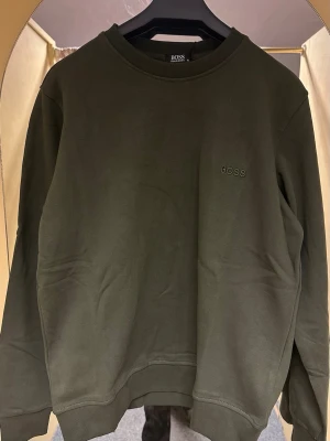 Mörkgrön sweatshirt från BOSS - Snygg mörkgrön sweatshirt från BOSS med diskret broderad logga på bröstet. Tröjan har rund hals, långa ärmar och är tillverkad i mjuk bomull. Perfekt för dig som gillar stilrena och enkla plagg med premiumkänsla.