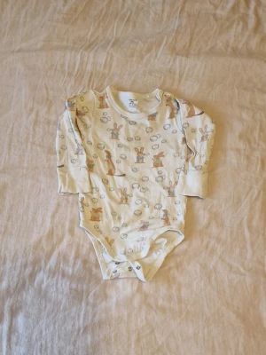 Vit body med djurmönster från Lindex - Supersöt vit body från Lindex med långa ärmar och tryckknappar i grenen. Mönstret består av kaniner och andra djur som blåser såpbubblor i beige, brun och blå toner. Tillverkad i mjuk bomull, perfekt för små äventyr. Det är ett litet streck på bodyns framsida som inte gick bort i vanlig tvätt (se bild).