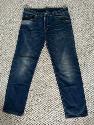Mörkblå jeans från Dsquared2 - Säljer ett par mörkblå jeans från Dsquared2 med klassisk femficksdesign och vit logotyp på bakfickan. Jeansen har en rak passform och snygga slitningar på framsidan för en cool look. Materialet är robust denim i bomull. Byxorna är köpta på Kids Brand Store