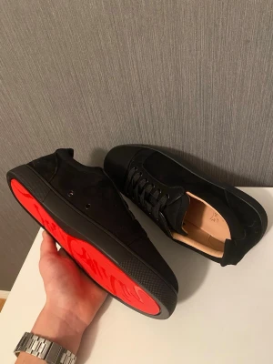 Christian Louboutin svarta sneakers - Säljer ett par svarta sneakers från Christian Louboutin med ikonisk röd sula och snygg snörning. Skorna är tillverkade i mocka och skinn, har rund tå och platt sula. Perfekt för dig som vill sticka ut med exklusiv design och lyxig känsla.