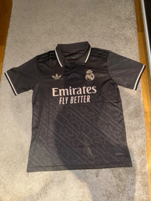 Adidas Real Madrid svart matchtröja S - Svart Real Madrid matchtröja från Adidas i storlek S. Tröjan har diskret mönster, broderade loggor och texten 'Emirates Fly Better' på bröstet. Klassisk krage med vita detaljer och AEROREADY-material som andas. Slim fit-modell, perfekt för fotboll eller träning.