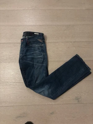 Replay Waitom jeans blå denim - Säljer ett par Replay Waitom jeans i klassisk blå denim med snygga slitningar och kontrastsömmar. Modellen har raka ben och normal passform, med detaljer som broderad logga och patch på bakfickan. Perfekt för dig som gillar stilrena jeans med lite edge.