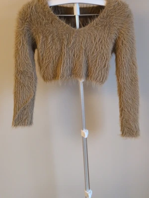 Beige fluffig cropped stickad tröja - Supersnygg beige stickad tröja med fluffig, lurvig textur och croppad passform. Tröjan har långa ärmar och bred urringning, perfekt för dig som vill ha en trendig och mysig look. Materialet känns mjukt och cozy mot huden.