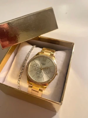 Guldig klocka från Regal med strass / metall guld/vit - Snygg guldig klocka från Regal med länkarmband och glittrande strass runt urtavlan. Klockan har en stilren design med tre små urtavlor.