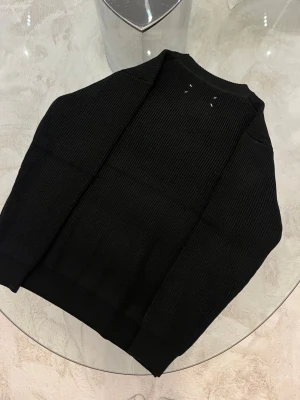Svart stickad tröja Maison Margiela - Säljer en svart ribbstickad tröja från Maison Margiela med klassisk rund halsringning och diskreta vita sömmar i nacken. Tröjan har en clean look och är tillverkad i ett mjukt, tjockt material som passar perfekt till kyligare dagar.
