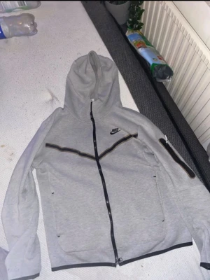 Grå zip hoodie från Nike - Säljer en grå hoodie från Nike med hel dragkedja och svart Nike-logga på bröstet. Hoodien har svarta detaljer längs dragkedjan och över bröstet samt en extra ficka med dragkedja på ena ärmen. Tillverkad i mjukt bomullsmaterial och har två sidofickor.