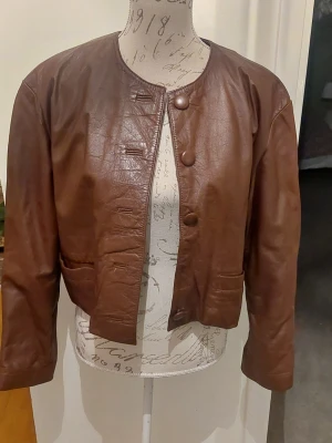 Brun kort skinnjacka utan krage - Snygg brun skinnjacka i kort modell med rund halsringning och stora knappar framtill. Jackan har två fickor och raka ärmar, 1 knapp har lossnat men ligger i fickan.