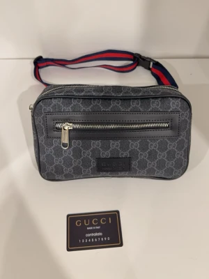 Gucci shoppingväska med monogram - Snygg shoppingväska från Gucci i mörkgrått med klassiskt GG-monogram över hela väskan. Väskan har en justerbar axelrem i rött och blått, silverfärgade dragkedjor och detaljer samt en praktisk ytterficka. Tillverkad i canvas med läderdetaljer.
