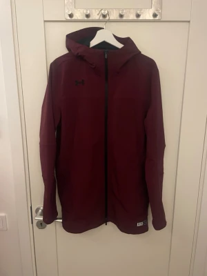Vinröd vindjacka från Under Armour - Snygg vinröd vindjacka från Under Armour med huva och hel dragkedja framtill. Jackan har minimalistisk design, diskret logga på bröstet och är tillverkad i ett lätt, vindtätt material. Perfekt för blåsiga dagar och enkel att matcha med olika outfits. 