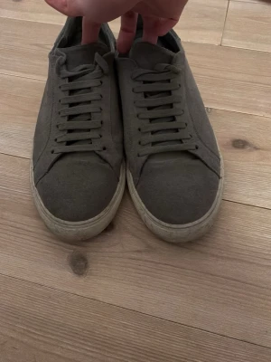 Gråa sneakers i mocka från Arigato - Stilrena gråa sneakers från Arigato i mjuk mocka med matchande grå skosnören och vit sula. Diskret guldfärgad logga på sidan. Klassisk låg modell som passar till det mesta och ger en clean look.