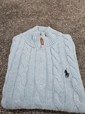Ljusblå kabelstickad tröja Polo Ralph Lauren - Ljusblå kabelstickad tröja från Polo Ralph Lauren med halv dragkedja och ribbad krage. Tröjan har det klassiska broderade logotypen på bröstet och är tillverkad i mjukt material. Perfekt för en stilren och avslappnad look.