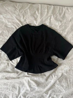 Svart figursydd  t-shirt från Gina Tricot - Snygg svart figursydd t-shirt från Gina Tricot med korta ärmar och markerad midja. Tillverkad i mjuk bomull som känns skön mot huden. Perfekt till jeans eller kjol för en trendig look.