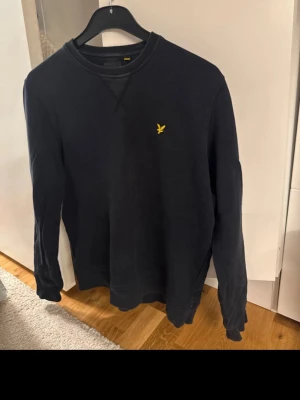 Svart sweatshirt från Lyle & Scott - Svart sweatshirt från Lyle & Scott med klassisk rund hals och ribbade muddar. Tröjan har ett broderat gult fågelmärke på bröstet och är tillverkad i mjuk bomull. Perfekt för chill dagar och enkel att matcha med jeans eller joggers.